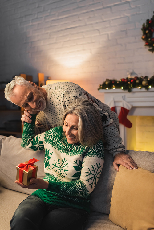 pleased middle aged woman holding christmas present and hugging husband in festive sweater