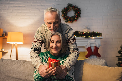 happy middle aged couple in festive sweaters holding christmas present in hands
