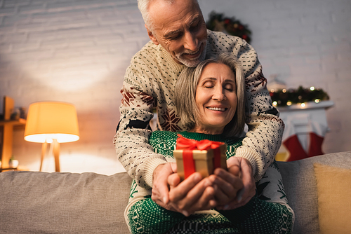 pleased middle aged couple in festive sweaters holding christmas present in hands