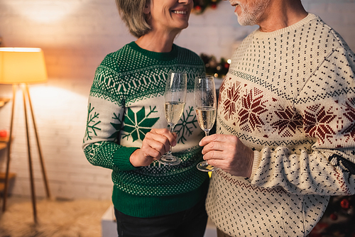 cropped view of happy middle aged couple in festive sweaters clinking glasses of champagne