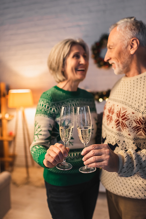 blurred and happy middle aged couple in festive sweaters clinking glasses of champagne