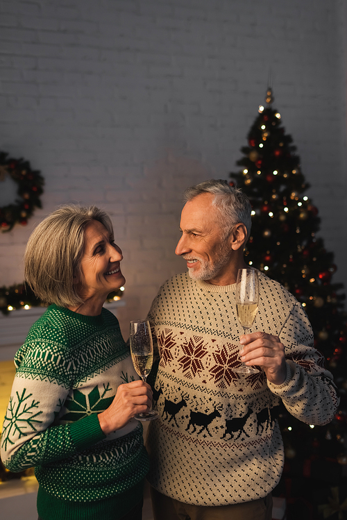 happy middle aged couple in festive sweaters holding glasses of champagne near christmas tree