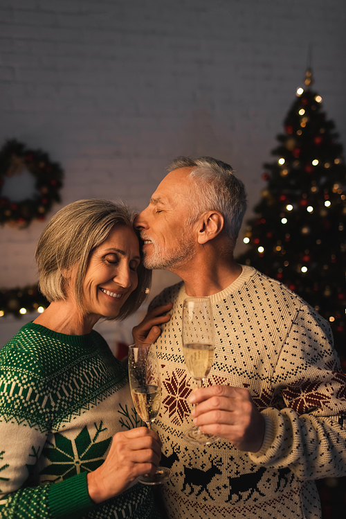 middle aged man in festive sweater holding glass of champagne and kissing happy wife near christmas tree