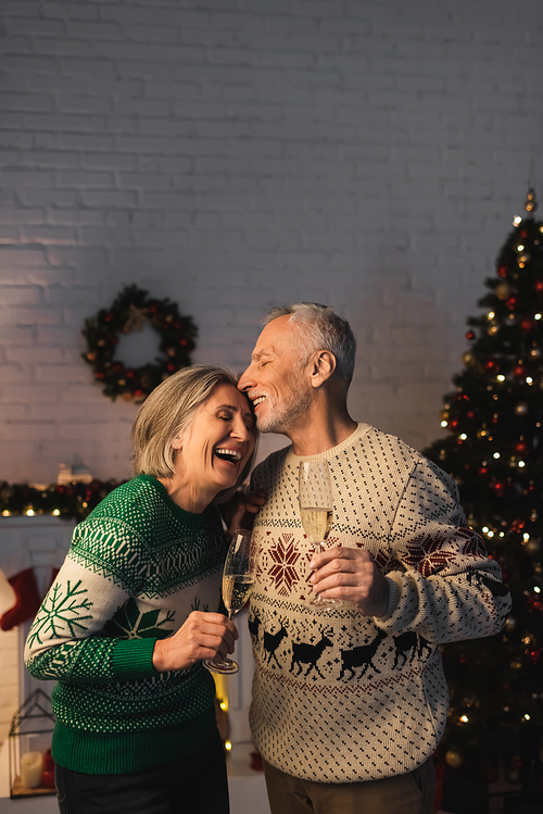 middle aged man in festive sweater holding glass of champagne and kissing joyful wife near christmas tree