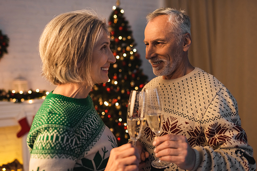 cheerful middle aged couple in festive sweaters clinking glasses of champagne near blurred christmas tree