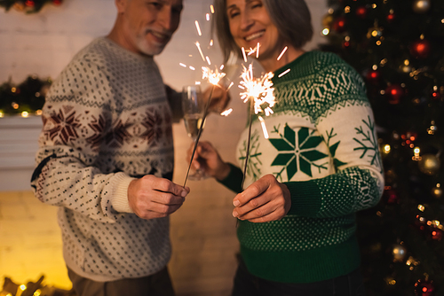 cheerful middle aged couple in festive sweaters holding bright sparklers near blurred christmas tree