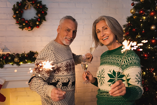 cheerful middle aged couple in festive sweaters holding bright sparklers and glasses of champagne on christmas eve