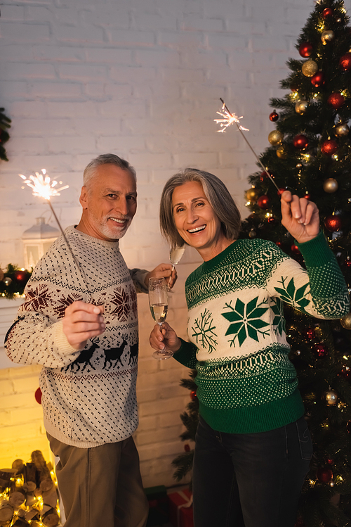 joyful middle aged couple in festive sweaters holding sparklers and glasses of champagne on christmas eve