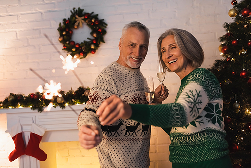 happy mature couple in festive sweaters looking at shiny sparklers and holding glasses of champagne on christmas eve