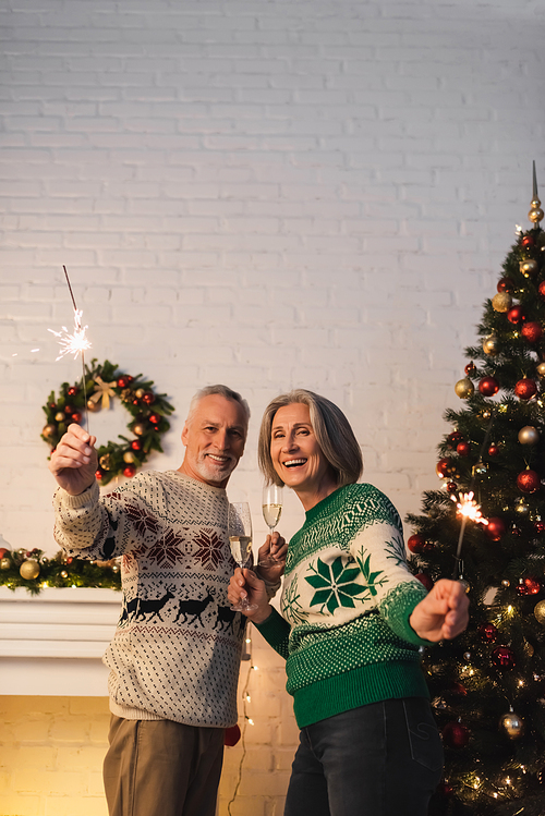happy mature couple in sweaters holding glasses of champagne and sparklers on christmas eve