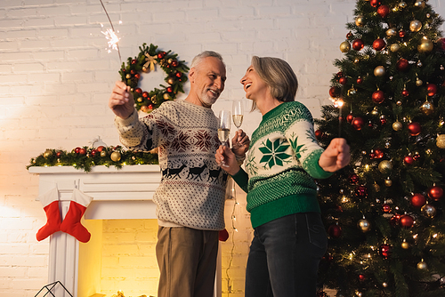 positive mature couple in sweaters holding glasses of champagne and sparklers on christmas eve