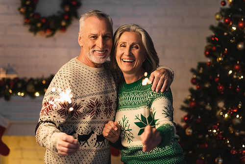 happy mature couple in holiday sweaters holding shiny sparklers on christmas eve at home