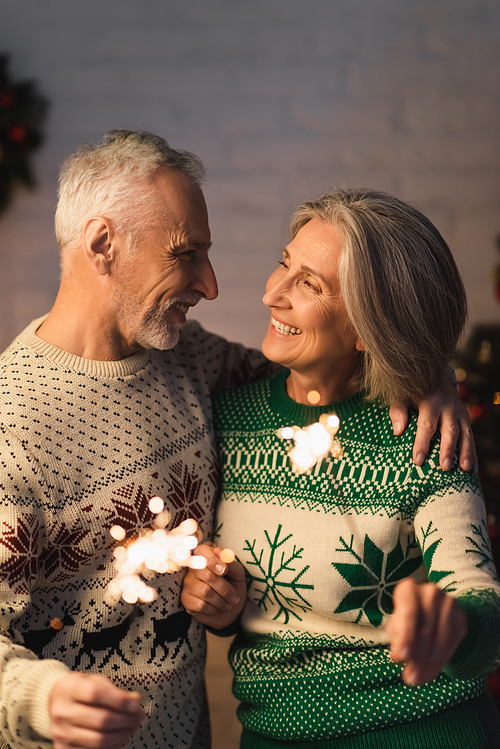 cheerful middle aged man hugging wife while holding sparkler on christmas eve
