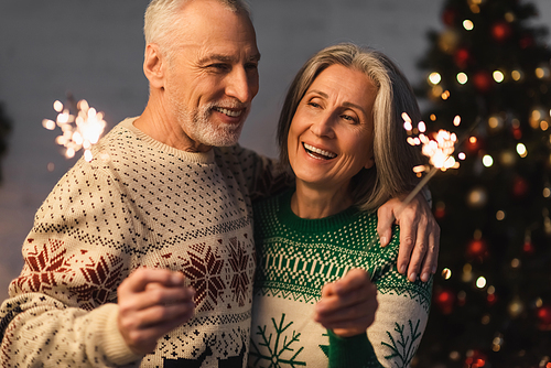 positive middle aged man hugging wife while holding sparkler on christmas eve