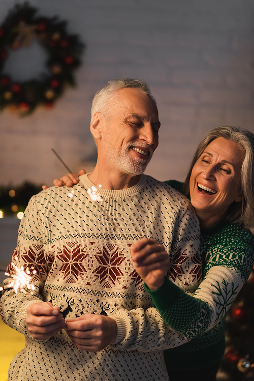 positive middle aged woman hugging bearded husband while holding sparkler on christmas eve