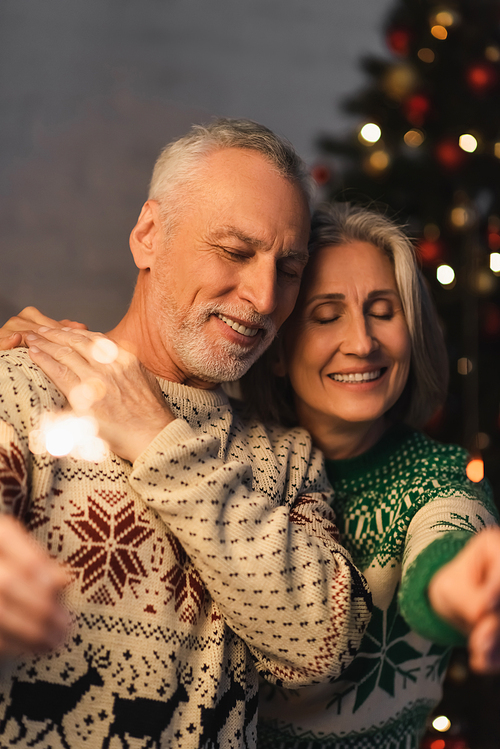 positive middle aged woman hugging bearded husband with closed eyes while holding sparkler on christmas eve