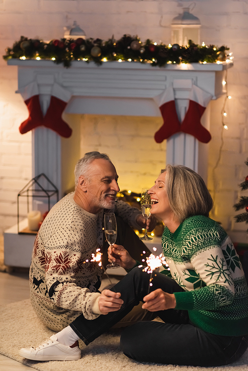 joyful middle aged man holding glass of champagne and looking at cheerful wife near shiny sparklers on christmas eve