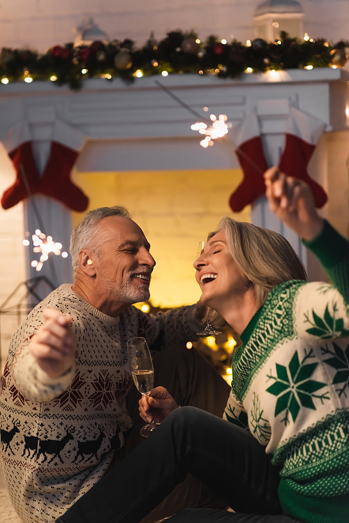 cheerful mature couple holding glass of champagne and shiny sparklers on christmas eve