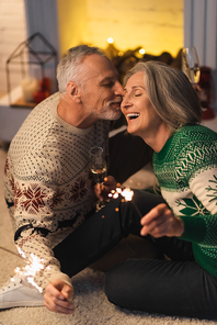 positive middle aged man kissing wife and holding shiny sparklers on christmas eve