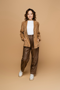 full length of brunette woman standing with hands in pockets of suede brown jacket on beige background