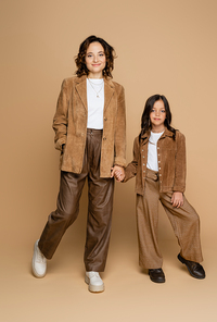 full length of mom and daughter in brown pants and suede jackets holding hands on beige background