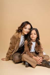 mother and daughter in suede jackets and brown pants smiling at camera while sitting on beige background