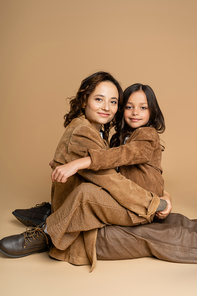 mother and daughter in suede jackets and brown pants smiling at camera while sitting on beige background