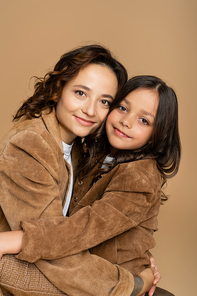 brunette mother and daughter in brown jackets hugging and smiling at camera isolated on beige