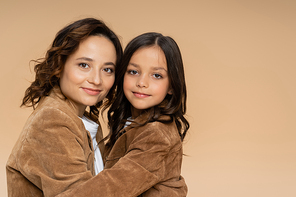 mom and daughter in stylish suede jackets smiling at camera while hugging isolated on beige