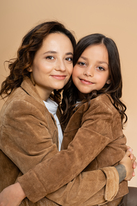 brunette mom and daughter in brown autumn jackets hugging and smiling isolated on beige