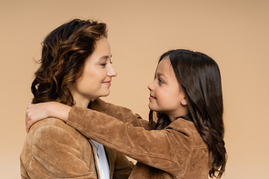 side view of mother and daughter in suede jackets smiling at each other isolated on beige