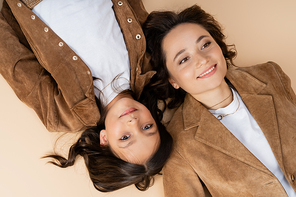 top view of brunette girl lying near mom in trendy autumn jacket and smiling at camera on beige background