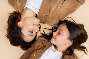 top view of mom and daughter in trendy autumn jackets smiling at each other on beige background