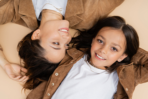 top view of happy and stylish woman looking at daughter smiling at camera and lying on beige background