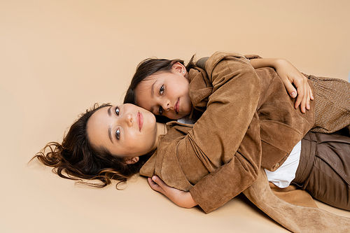 positive mom and daughter in trendy autumn jackets lying and looking at camera on beige background