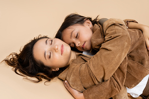 mother and child in stylish suede jackets lying with closed eyes and hugging on beige background