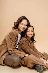 positive mother and child in suede jackets and brown pants sitting and looking at camera on beige background