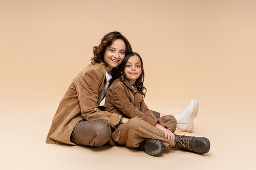 happy woman and girl in trendy boots and brown suede jackets sitting on beige background