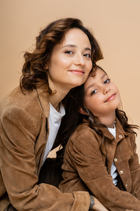 brunette woman and girl in stylish autumn jackets smiling at camera isolated on beige