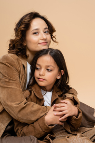 happy and stylish woman embracing daughter in brown suede jacket isolated on beige