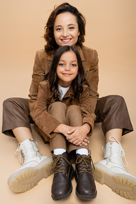 happy woman and kid in suede jackets and trendy boots sitting on beige background