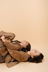 side view of girl having fun and lying on happy mom in autumn clothes on beige background
