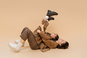 side view of girl with raised legs having fun and lying on mom in autumn outfit on beige background