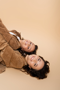 top view of cheerful woman and child in brown suede jackets lying and looking at camera on beige background