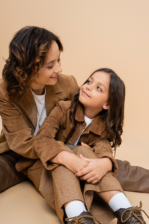 joyful woman and child in stylish suede jackets looking at each other on beige background