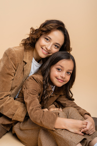 pleased mother and daughter in brown suede jackets looking at camera on beige background