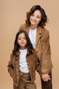 cheerful woman holding hands with trendy daughter posing isolated on beige