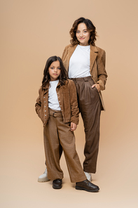 full length of woman and girl in trendy autumn clothes posing with hands in pockets on beige background