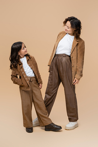 cheerful mom and daughter in suede jackets and trendy pants looking at each other on beige background
