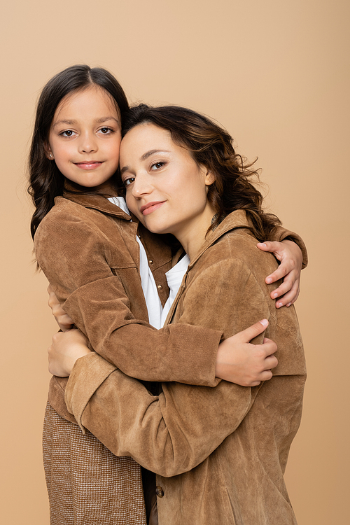young woman and girl in stylish autumn jackets embracing and smiling at camera isolated on beige